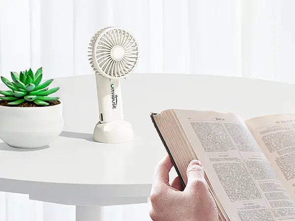 Mini ventola usb bianca su tavolo bianco accanto a pianta verde e persona che legge un libro
