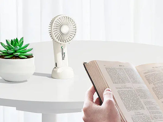 Mini ventola usb bianca su tavolo bianco accanto a pianta verde e persona che legge un libro